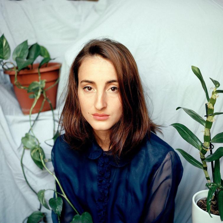 Photographie en couleur d'une jeune femme châtain aux cheveux mi-longs, avec des plantes autour d'elle. 